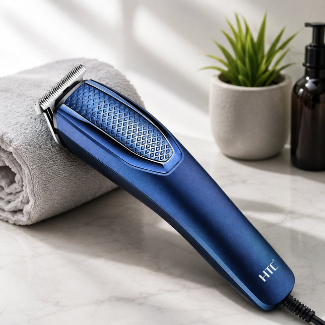 A blue HTC cordless hair clipper resting on a gray towel on a white marble surface, with a small potted plant and a dark bottle in the background.