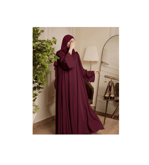Woman wearing a burgundy abaya in a room with a lamp and plant.