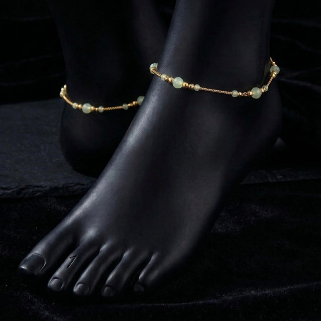 Gold anklets with green beads on a black foot mannequin against a dark background